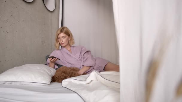 Jovem mulher usando telefone na cama deitada com cão no quarto minimalista — Vídeo de Stock