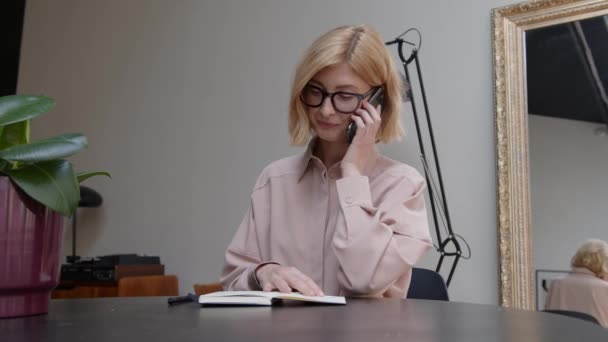 Blondine vrouw praten aan de telefoon schrijven in notebook op de tafel — Stockvideo