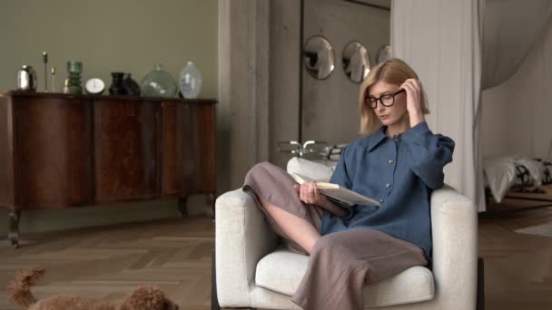 一个带着书的金发女人坐在扶手椅上，可爱的小狗 — 图库视频影像