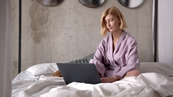 Blonde woman using laptop in bathrobe sitting on white linens — Stock Video