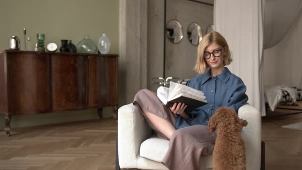 Femme blonde avec livre à la main assise dans un fauteuil, caressant un animal — Video