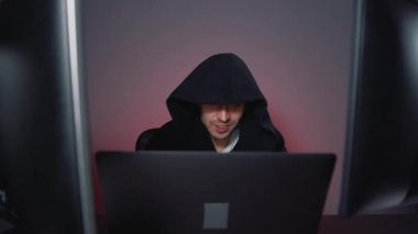 Siyah başlıklı adam, birkaç bilgisayarla çalışıyor. Şeytani gülümseme.