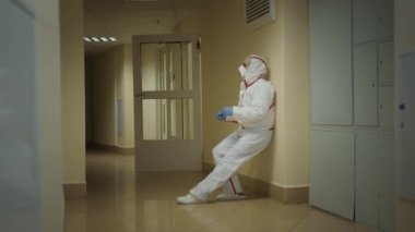 Tehlikeli madde giysisi giymiş yorgun bir doktor yere oturup koruyucu maskesini çıkarıyor.