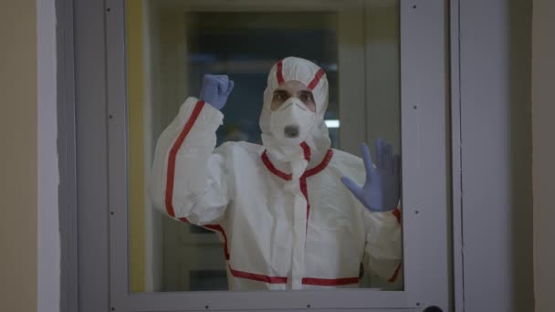 Doktor v ochranném obleku s maskou udeří do skla na dveřích v budově — Stock video