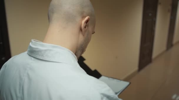Доктор у білому пальто, який дивиться через буфер обміну в коридорі в лікарні — стокове відео