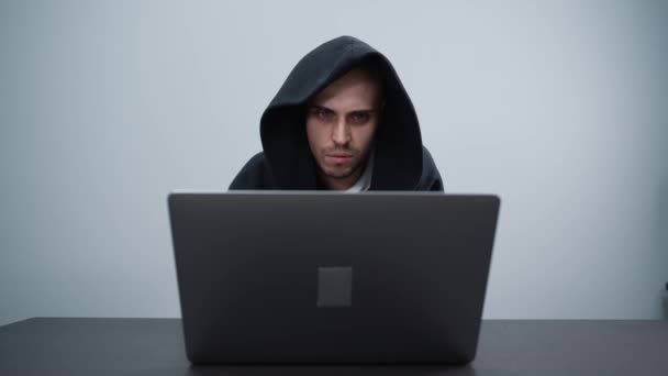 Нервовий хакер в чорному капюшоні на сірому фоні, що працює з ноутбуком — стокове відео