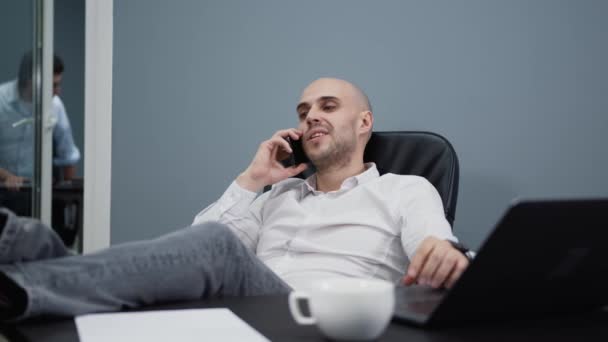 Homme de bureau détendu souriant, faisant un appel téléphonique avec les jambes sur la table — Video