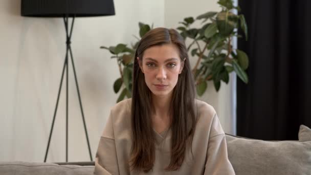 Брюнетка в бежевом свитере смотрит в камеру на современный домашний интерьер — стоковое видео