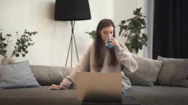 Esmer kadın dizüstü bilgisayar kullanıyor ve koltukta oturup bir bardak su içiyor.