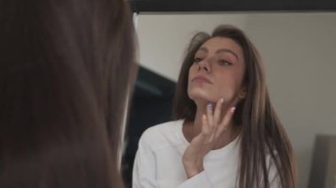 Genç bir kadın gülümsüyor, aynaya bakıyor, saçını düzeltiyor ve tenine dokunuyor.