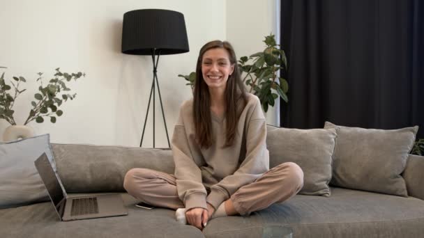 Mujer joven con portátil en un sofá, sonriendo y agitando las manos en el interior del hogar — Vídeos de Stock