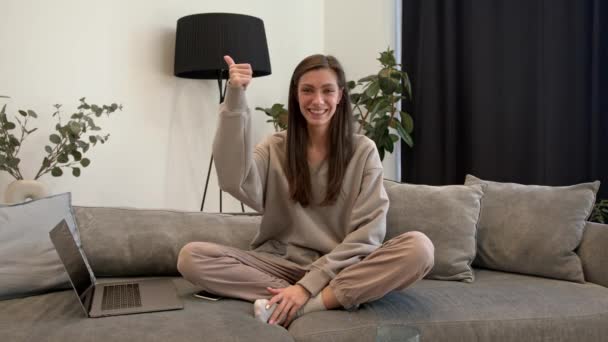 Молода жінка з ноутбуком на дивані, посміхаючись і показуючи великий палець вгору і знак миру — стокове відео