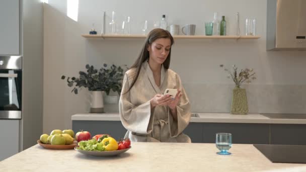 O femeie atractivă care trimite mesaje la telefon în mâini bea un pahar de apă în bucătărie — Videoclip de stoc