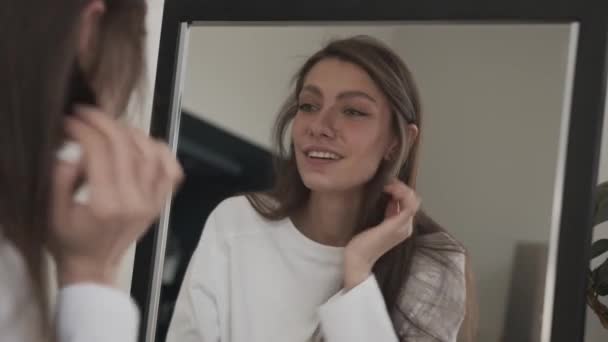 Mujer joven sonriendo, mirándose en el espejo, arreglando el pelo y tocándose la piel — Vídeos de Stock