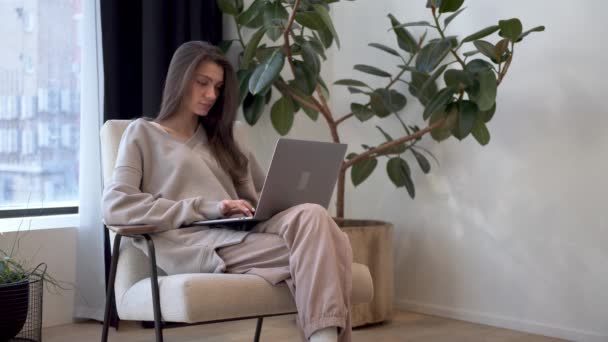 Fiatal nő hosszú haj dolgozik, ül karosszékben laptoppal ablak mellett — Stock videók