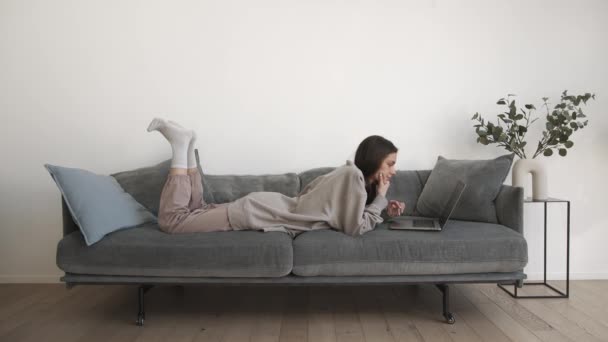 Молодая женщина фрилансер работает, лежа на сером диване в гостиной на дому — стоковое видео