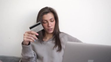 Elinde ve bilgisayarında kredi kartı olan bir kadın oturma odasındaki kanepeye yakın çekim yapıyor.