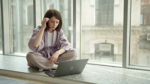 Brünette Frau tippt mit Laptop am Fenster auf der Fensterbank — Stockvideo