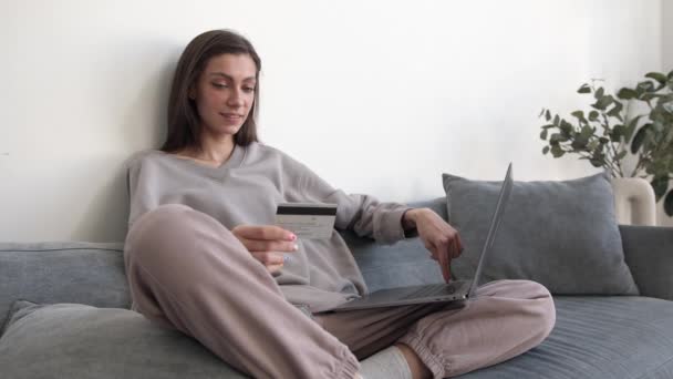 Jonge vrouw met creditcard in de hand en laptop zittend op de bank in de woonkamer — Stockvideo