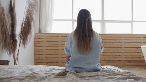 Ung kvinna sitter på kanten av sängen tittar på fönstret — Stockvideo