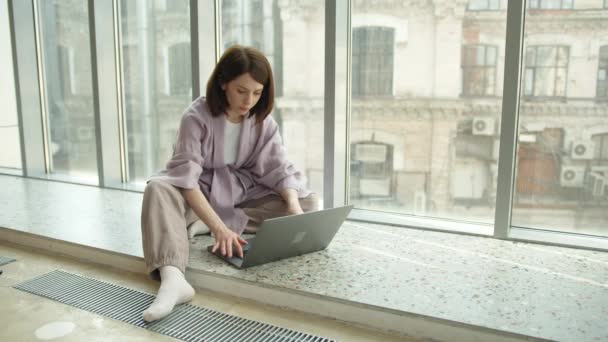 Businesswoman psaní pomocí notebooku sedí v blízkosti okna na parapetu — Stock video
