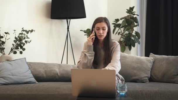 布鲁内特的女人坐在沙发上，拿着手提电脑在讲电话 — 图库视频影像