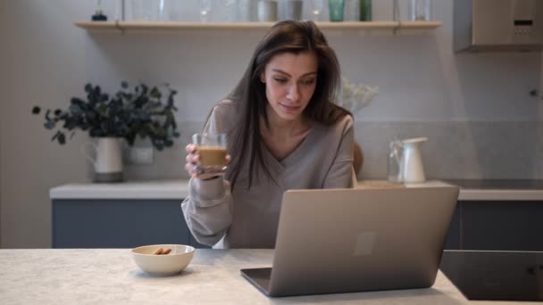 Młoda kobieta je śniadanie i wykonuje wideo połączenie w laptopie w kuchni — Wideo stockowe