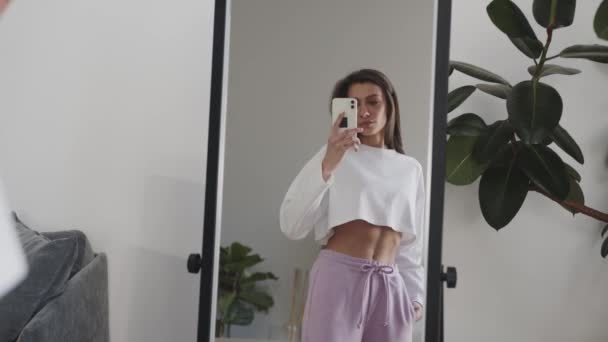 Молодая женщина, стоящая с телефоном перед зеркалом, делает фото — стоковое видео