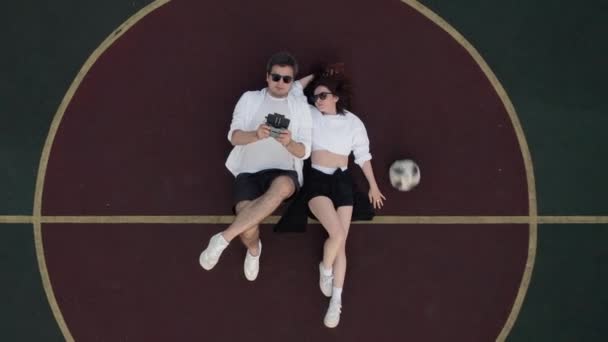 Une femme et un homme allongés sur un terrain de basket de rue en ville, un drone monte. — Video