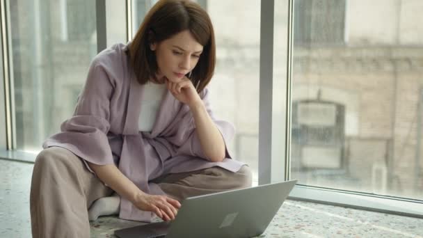 Businesswoman psaní pomocí notebooku sedí v blízkosti okna na parapetu — Stock video