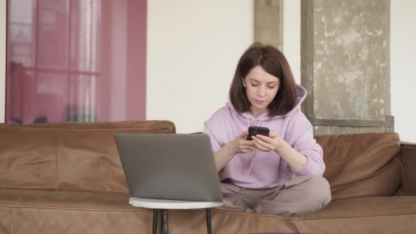 Жінка з Брюнета пише смс по телефону сидячи на дивані з ноутбуком. — стокове відео