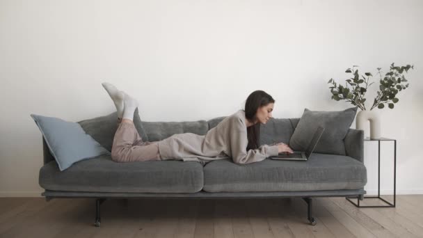 Mujer joven freelancer trabajando, tumbada en un sofá gris en la sala de estar en casa — Vídeos de Stock