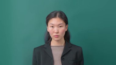 Siyah ceketli ve kahverengi gömlekli ciddi bir genç kadın yeşil stüdyo arkaplanına karşı duruyor. Profesyonellik ve kendine güven kavramı.