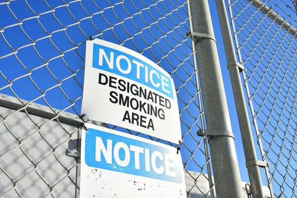 An image of a  blue and white designated smoking area sign posted to a chain link fence.