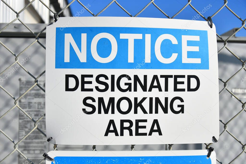 An image of a  blue and white designated smoking area sign posted to a chain link fence.