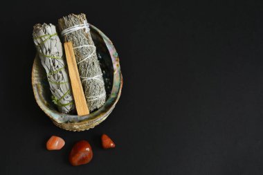 Siyah zemin üzerinde çakra iyileştirici kristaller bulunan deniz kulağı kabuğundaki beyaz adaçayı lekesi demetlerinin üst görünümü.