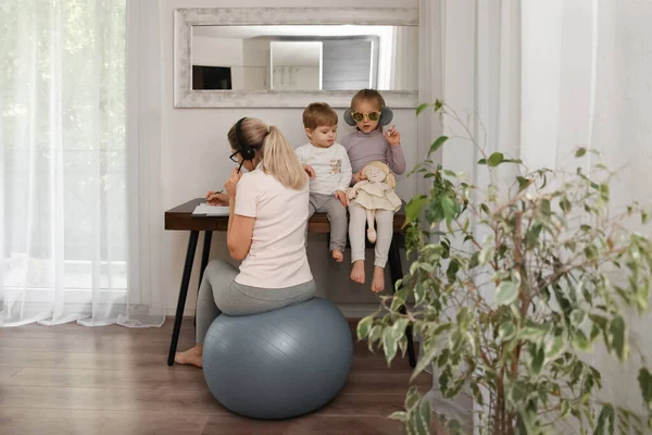 Mother sits on a fitness ball in headphones and works at home with her children during quarantine Covid-19