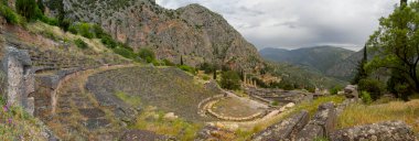 Panoramic view on ancient theater and temple of Apollo with columns in Delphi against the background of mountains and the sky with clouds in Greece