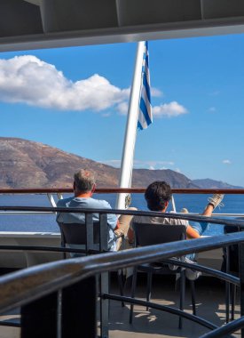 Andros, Yunanistan. 30 Eylül 2025: Yunanistan 'daki Tinos ve Andros adalarının arka planına karşı Ege Denizi' nde Yunan bayrağı altında bir feribotta bir baba ve oğul