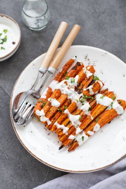 Kızarmış havuç çubukları beyaz bir tabakta. Gri arka planda otlu ve baharatlı fırında havuç. Lezzetli vejetaryen yemeği. Sağlıklı kök sebze kavramı