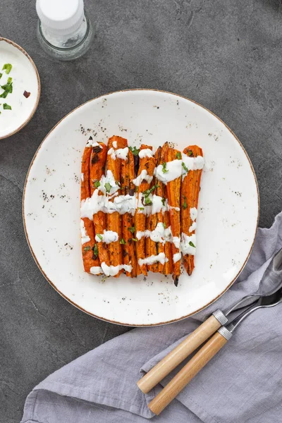 Kızarmış havuç çubukları beyaz bir tabakta. Gri arka planda otlu ve baharatlı fırında havuç. Lezzetli vejetaryen yemeği. Sağlıklı kök sebze kavramı