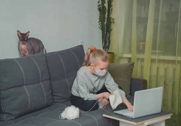 Beyaz kız örgü örmeyi öğreniyor, karantinadaki dizüstü bilgisayar derslerinde eğitim videoları izliyor.