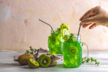 Bir kadın cam kavanoz kivi suyu ya da smoothie üzerinde pipet içerek elini tutuyor. Kiwi Mojito kokteyli ya da alkolsüz mocktail naneli ve dilimlenmiş kivi meyveli ahşap arka planda fotokopi alanı