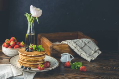Çilekli krep, seramik tabakta yaban mersini ve nane şekeri, küçük seramik kavanozdan şurup ve koyu ahşap bir masada çiçekler.