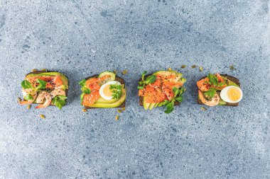 Sağlıklı atıştırmalık ya da somon, avokado, karides, yumurta, salatalık, kuzu salatası ya da mısır salatası ve maydanoz ile aperatif tost. Açık gri masa yüzeyi