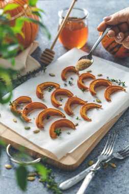 Kadının eli kahverengi şekeri fırında pişmiş mini balkabağı dilimlerinin üzerine kekik serpiyor. Gri masanın üzerindeki tahta tahtada kekik var. Mevsimlik vejetaryen yemek tarifi