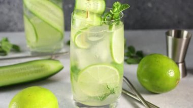 Mojito kokteyli, nane, salatalık ve kireç karışımını gri beton yüzey arka planında viski bardağında süslemek. Kamera cam boyunca hareket ediyor.