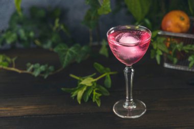 Koyu ahşap masa yüzeyinde büyük bir buz yumağı bulunan zarif kadehteki kan portakallı kokteyl sepetinde yeşil otlar ve çiğ taze kan portakalı vardı..