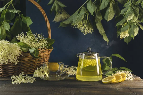 Herbal health tea of elder flower surrounded fresh flowers and leaves in basket, dark rustic style. Close up.