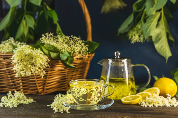 Herbal health tea of elder flower surrounded fresh flowers and leaves in basket, dark rustic style. Close up.